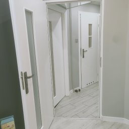 Profeska usługi malarskie Krzysztof Kuczyński - Nowoczesne, białe drzwi wewnętrzne z przeszkleniem, widok na kolejne drzwi w głębi mieszkania, szara ściana, jasna podłoga imitująca drewno, widoczny fragment opakowania zabawki i czarna torba przy...