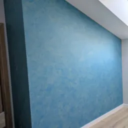 Błękitna ściana z efektem dekoracyjnym w wykończonym wnętrzu, widok na fragment poddasza z oknem dachowym i drewnianą podłogą.