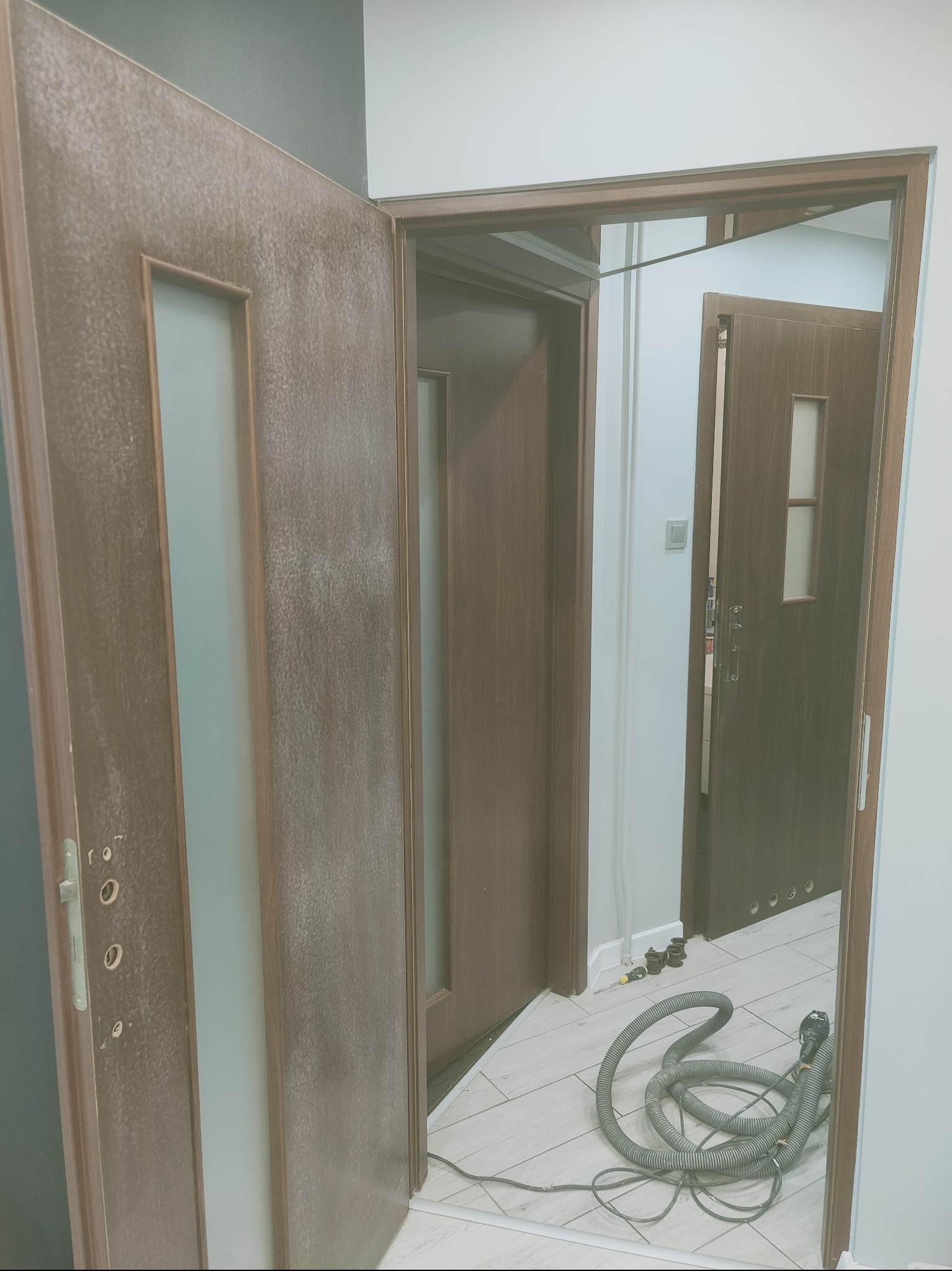 Dwa brązowe, częściowo otwarte drzwi z szybami, widoczne ślady szpachlowania lub malowania, na podłodze wąż od odkurzacza, widok na kolejne drzwi w tle.