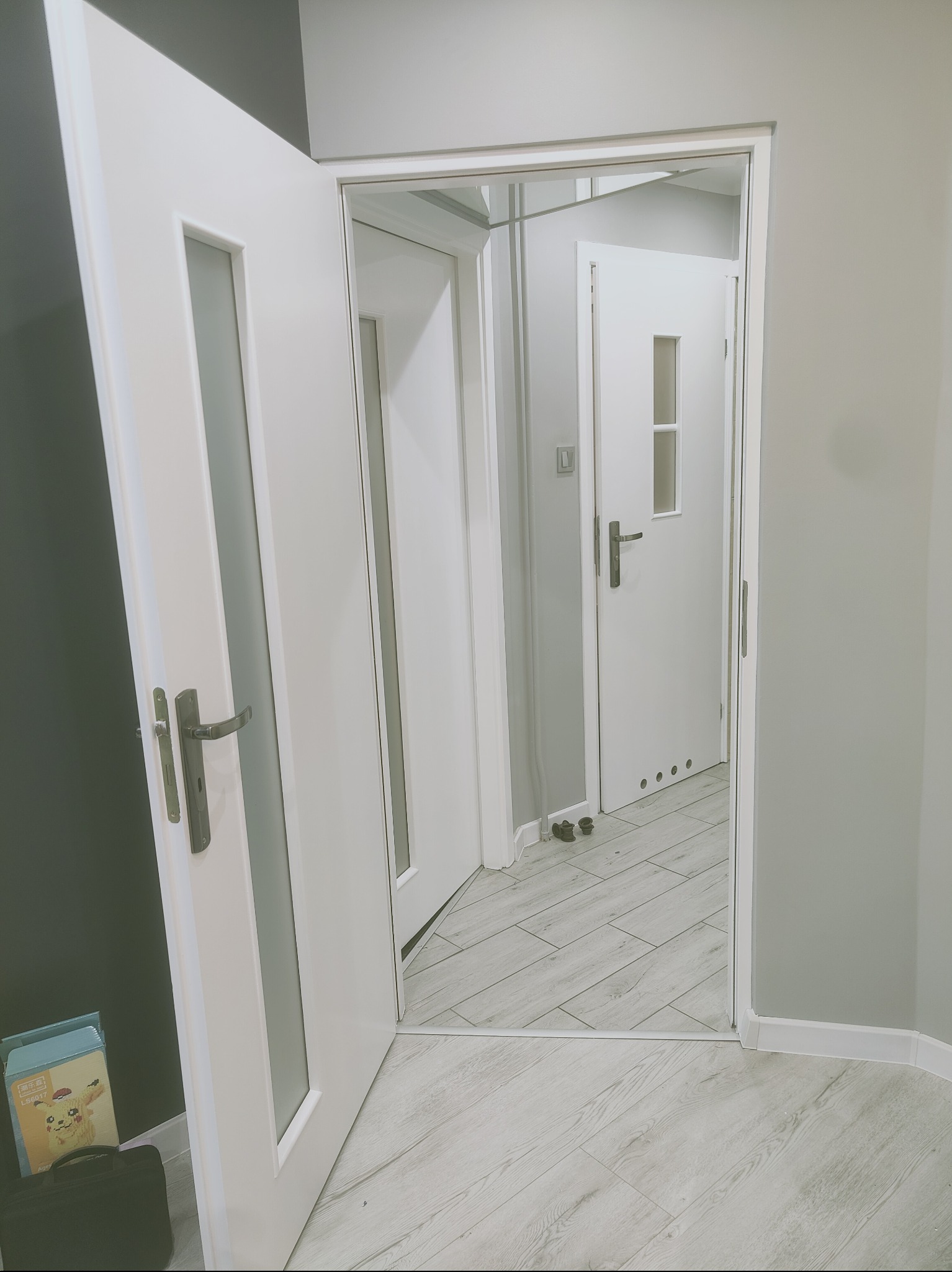 Nowoczesne, białe drzwi wewnętrzne z przeszkleniem, widok na kolejne drzwi w głębi mieszkania, szara ściana, jasna podłoga imitująca drewno, widoczny fragment opakowania zabawki i czarna torba przy...