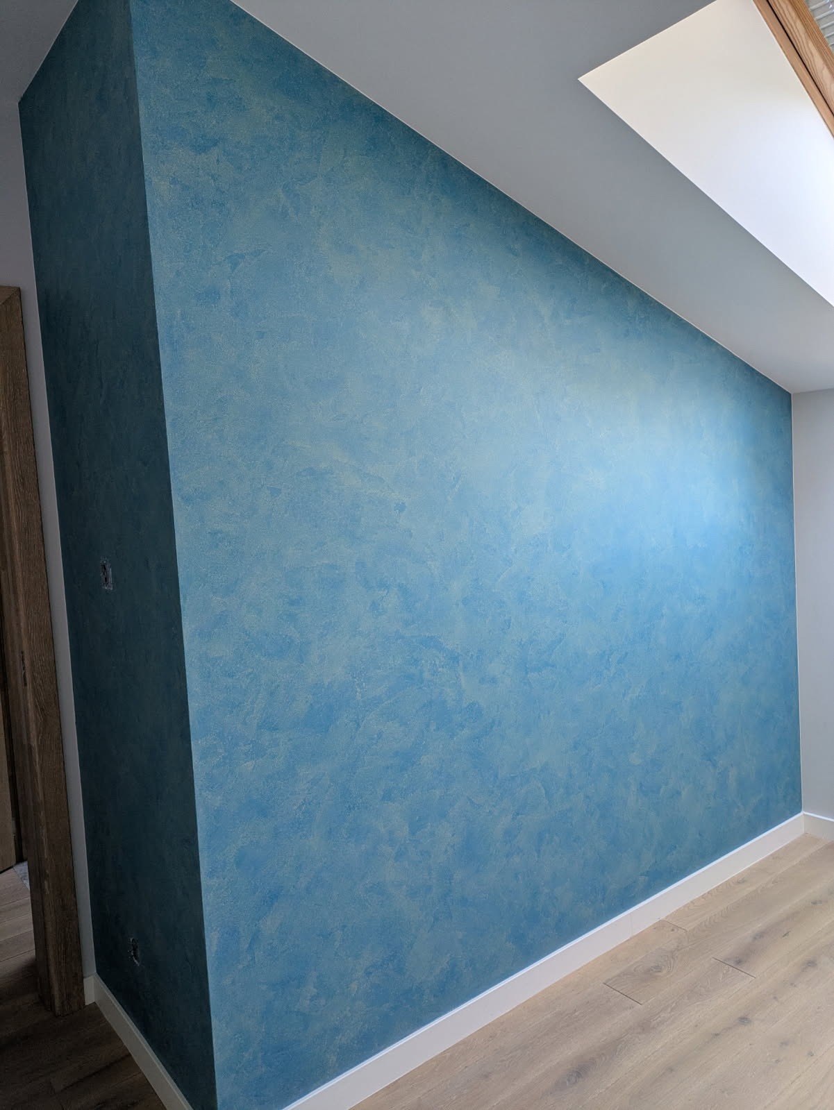 Błękitna ściana z efektem dekoracyjnym w wykończonym wnętrzu, widok na fragment poddasza z oknem dachowym i drewnianą podłogą.