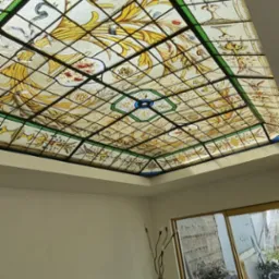 Sufit z dekoracyjnym witrażem w geometrycznej ramie, zdobiony motywami roślinnymi i ptaków, widok z dołu w jasnym pomieszczeniu z kanapą i roślinami.