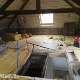 Poddasze w trakcie ocieplania: widoczne drewniane krokwie, okno dachowe, częściowo ułożona izolacja Isover pomiędzy legarami podłogowymi oraz instalacja elektryczna i hydrauliczna prowadzona...