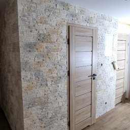 Wykończenia wnętrz - Wykończone wnętrze z kamienną okładziną ściany, drewnopodobną podłogą i zamontowanymi drzwiami wewnętrznymi z poziomymi frezami; widoczne puszki instalacyjne w suficie.