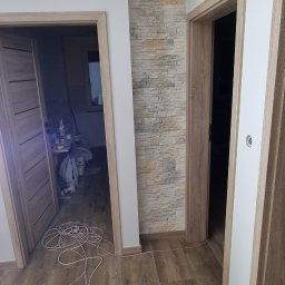 Wykończenia wnętrz - Wnętrze w trakcie remontu: położona podłoga z płytek imitujących drewno, widoczne trzy ościeżnice drzwiowe z zamontowanymi drzwiami po lewej, ściana wyłożona dekoracyjnym kamieniem, przedłużacz...