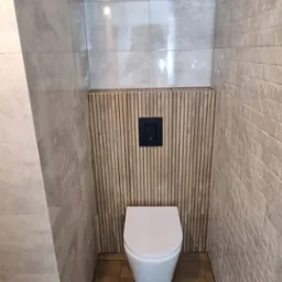 Nowoczesna toaleta podwieszana z czarnym przyciskiem spłukującym na tle drewnianych lameli, szara płytka imitująca beton na ścianach i podłodze, na podłodze leży taśma malarska.