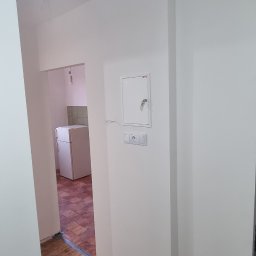 Wykończenia wnętrz - Świeżo pomalowane na biało ściany w mieszkaniu, widoczny fragment podłogi z drewnianych desek i wiadro z farbą. W tle widoczna kuchnia.