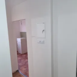 Świeżo pomalowane na biało ściany w mieszkaniu, widoczny fragment podłogi z drewnianych desek i wiadro z farbą. W tle widoczna kuchnia.