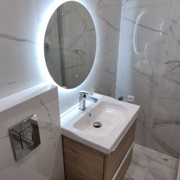 Wykończenia wnętrz - Nowoczesna łazienka z marmurowymi płytkami, okrągłym lustrem z podświetleniem LED, drewnianą szafką pod umywalką i podwieszaną toaletą. Minimalistyczny design.