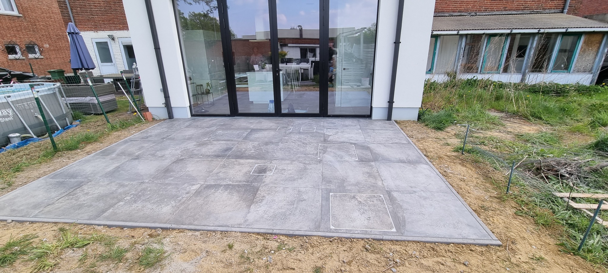 Szara, kwadratowa nawierzchnia tarasu z płytek, z widocznymi włazami rewizyjnymi, przylegająca do dużych, przeszklonych drzwi balkonowych w nowoczesnym domu.