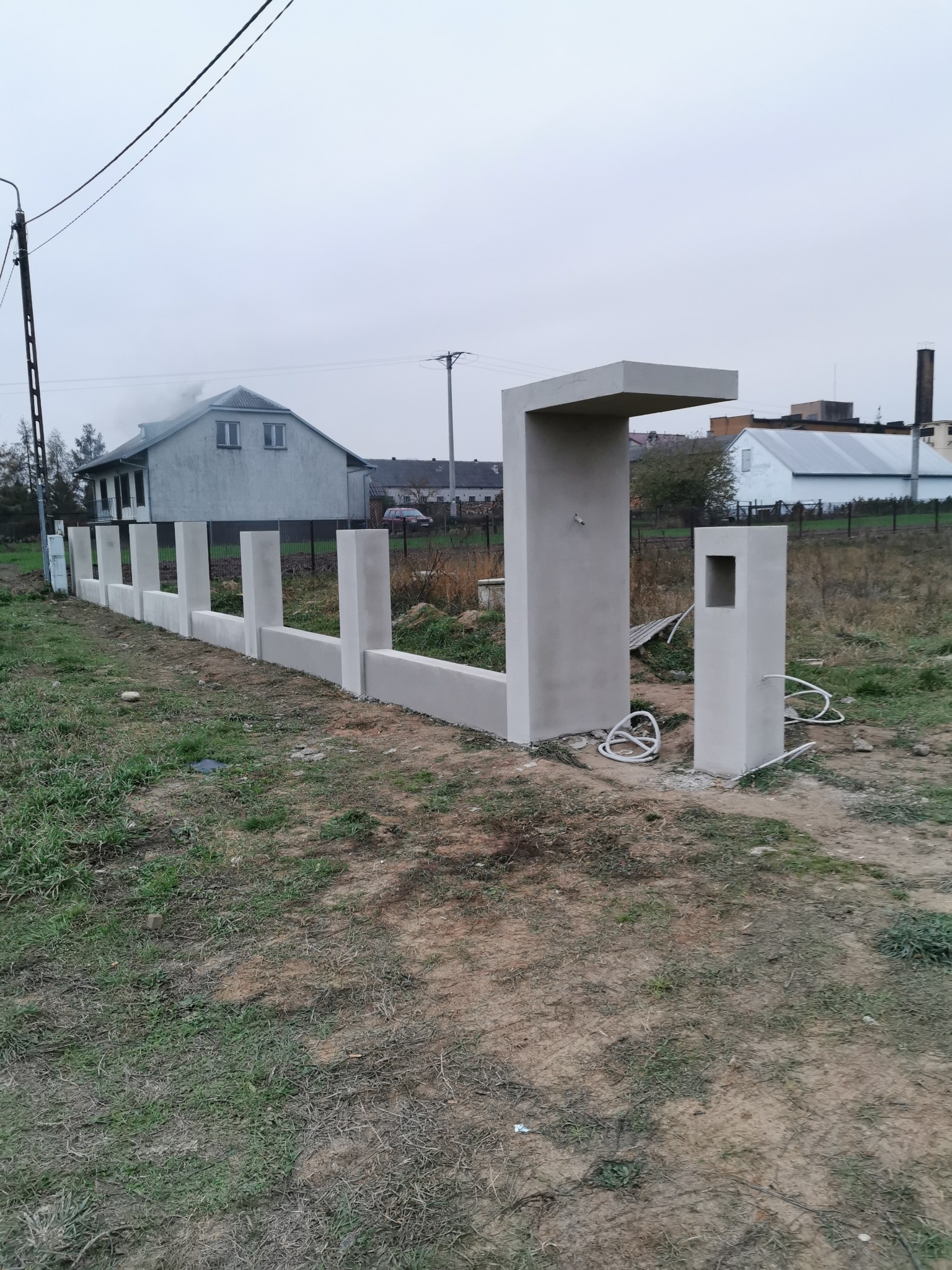 Surowy, szary, betonowy płot w trakcie budowy, z widocznymi przewodami i otworami, na tle domu i pochmurnego nieba.