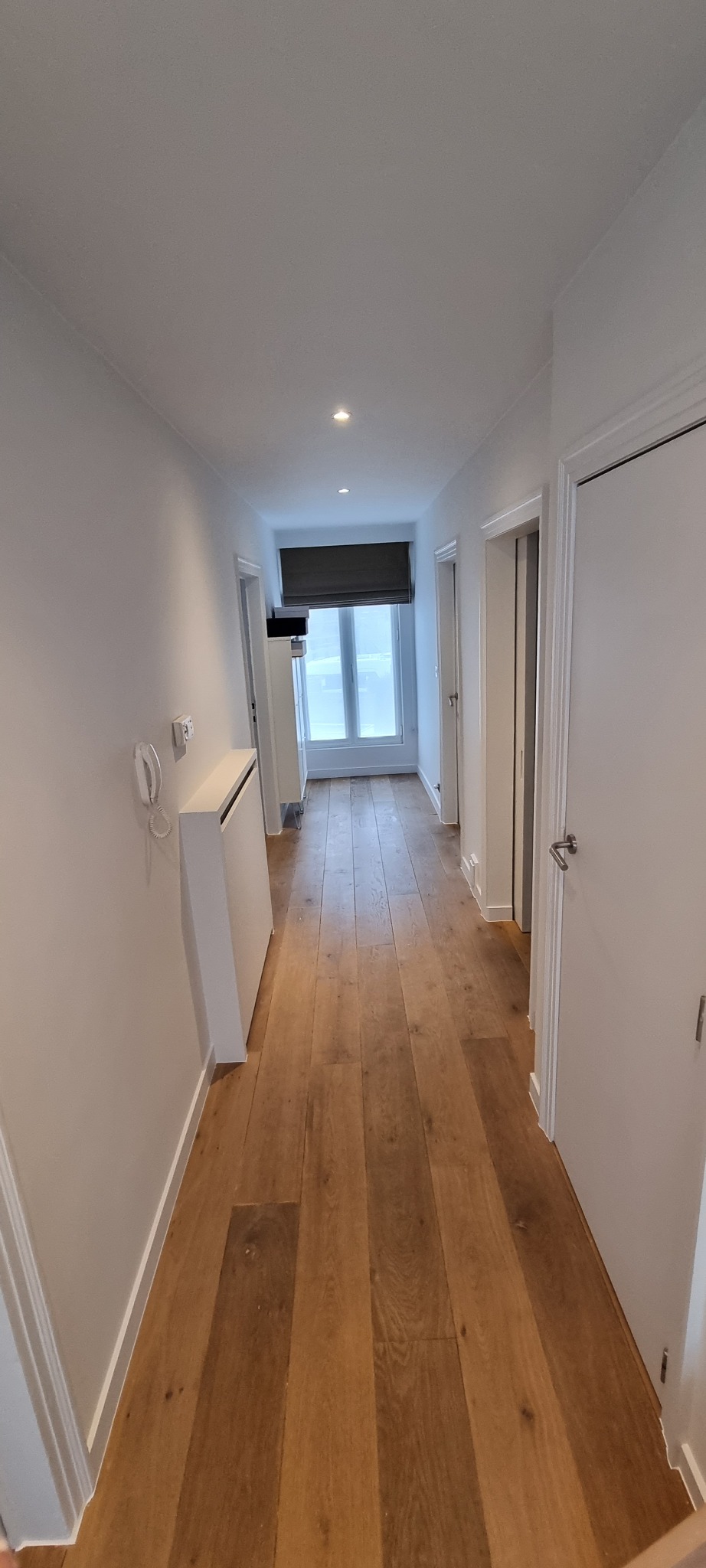 Długi korytarz z parkietem ułożonym w deski, białe ściany z grzejnikiem i telefonem na ścianie, okno zasłonięte roletą rzymską, kilka par drzwi po bokach.