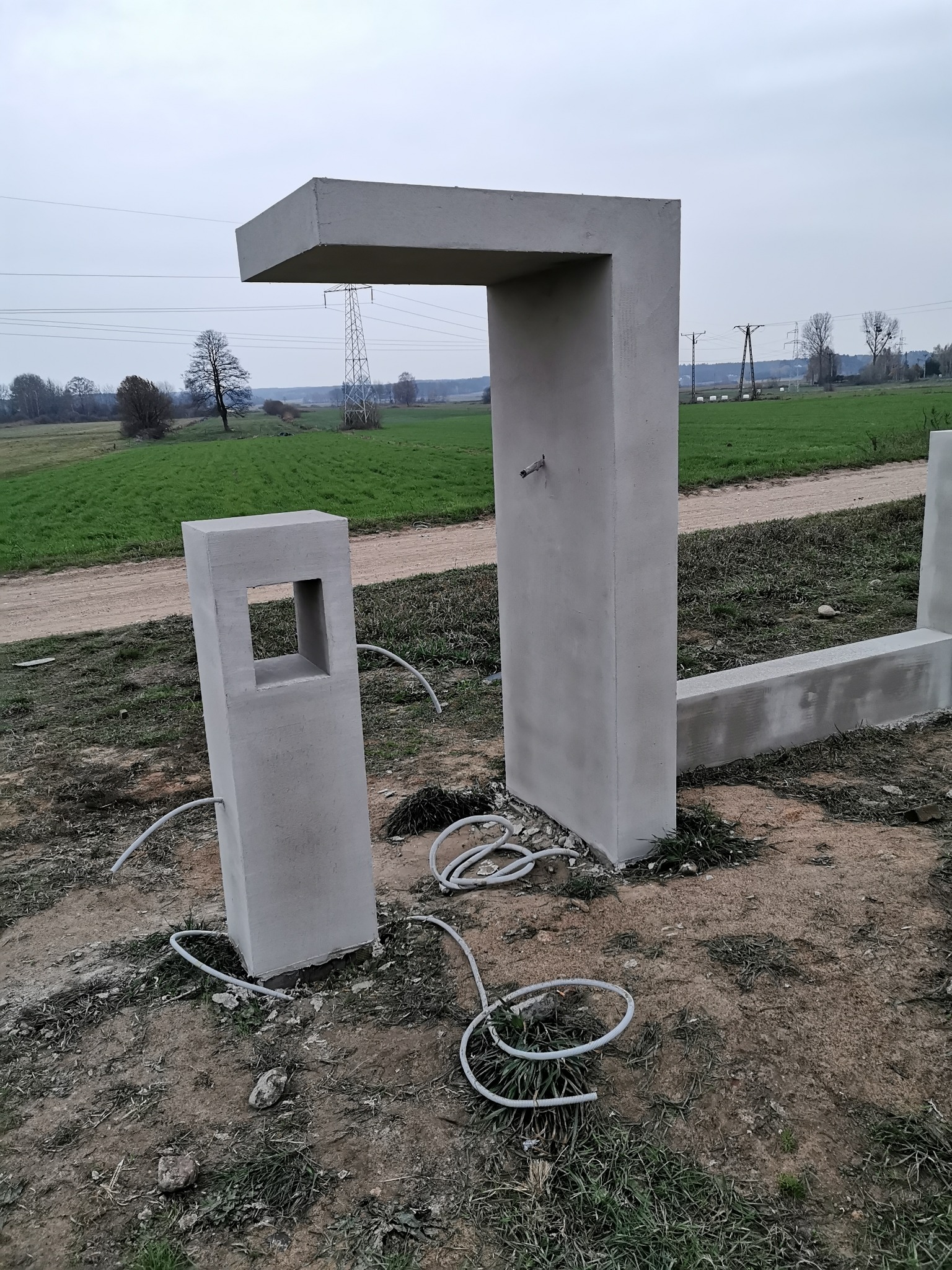 Surowa, betonowa konstrukcja w kształcie litery L z wystającym kranem i betonowy słupek z otworem, otoczone przewodami, na tle pola i słupów energetycznych.