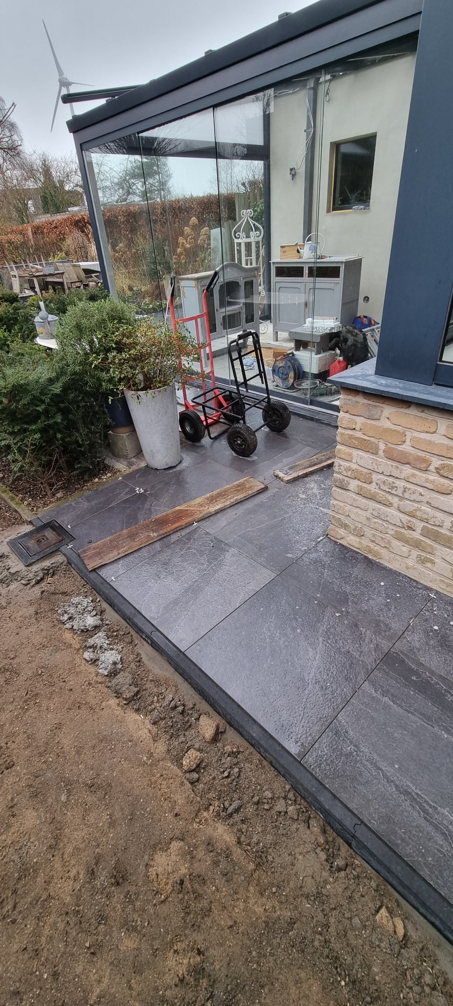 Ułożona, ciemnoszara posadzka z kamiennych płyt na zewnątrz budynku z przeszkloną werandą, widoczne narzędzia i donica z rośliną.