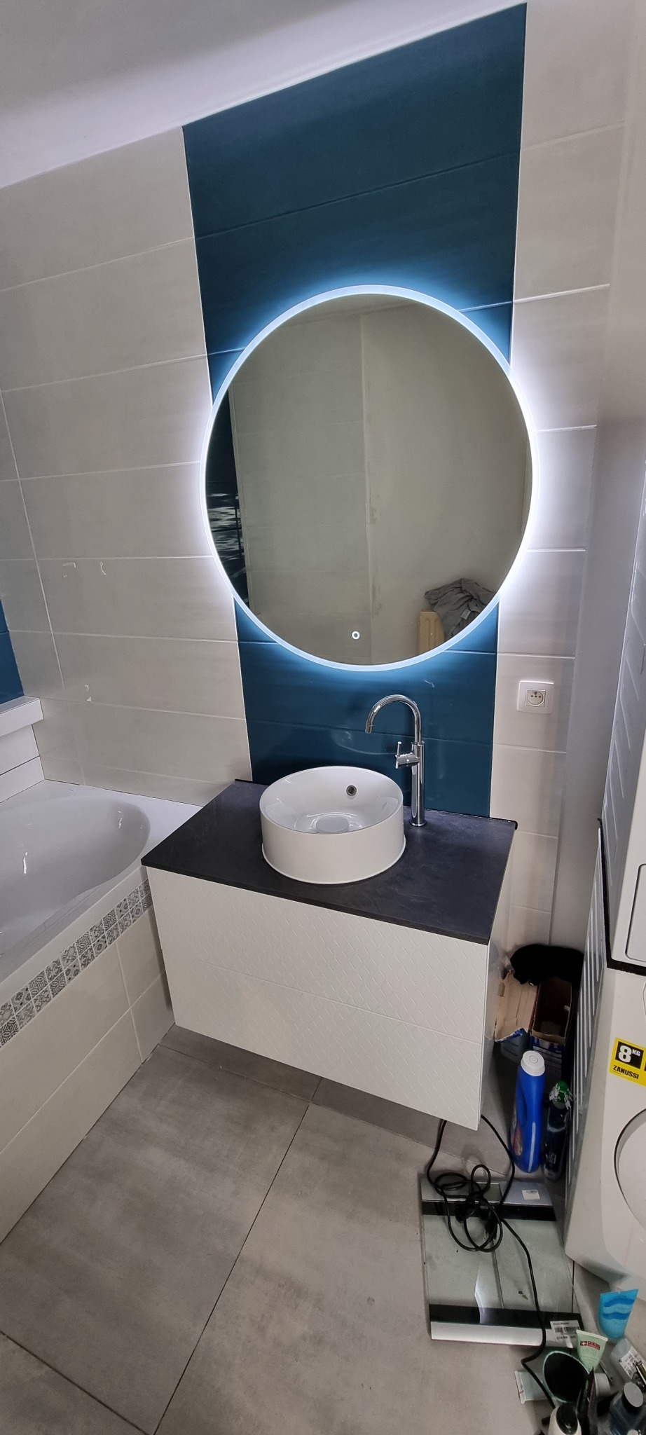 Nowoczesna łazienka z umywalką nablatową, okrągłym lustrem z podświetleniem LED, białymi i granatowymi płytkami oraz szarą podłogą. W tle widoczna wanna i pralka, a na podłodze waga łazienkowa.