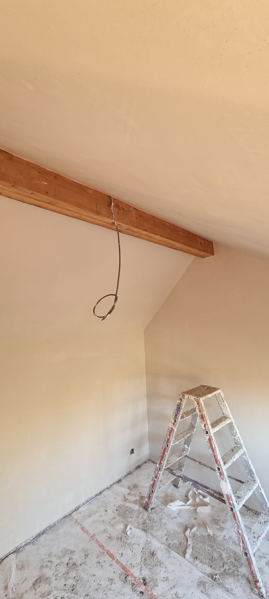 Poddasze po nałożeniu gładzi, widoczna drewniana belka stropowa, wiszący kabel elektryczny, aluminiowa drabina malarska i zabrudzona podłoga.