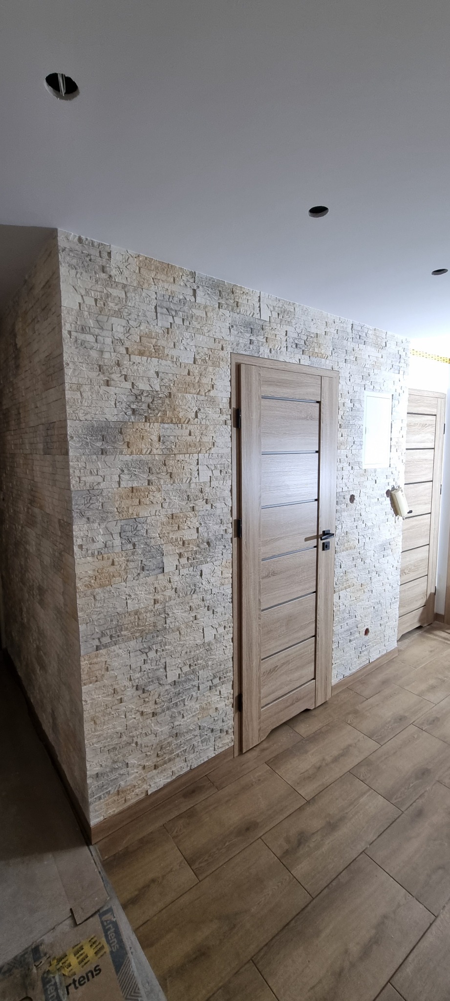 Wykończone wnętrze z kamienną okładziną ściany, drewnopodobną podłogą i zamontowanymi drzwiami wewnętrznymi z poziomymi frezami; widoczne puszki instalacyjne w suficie.