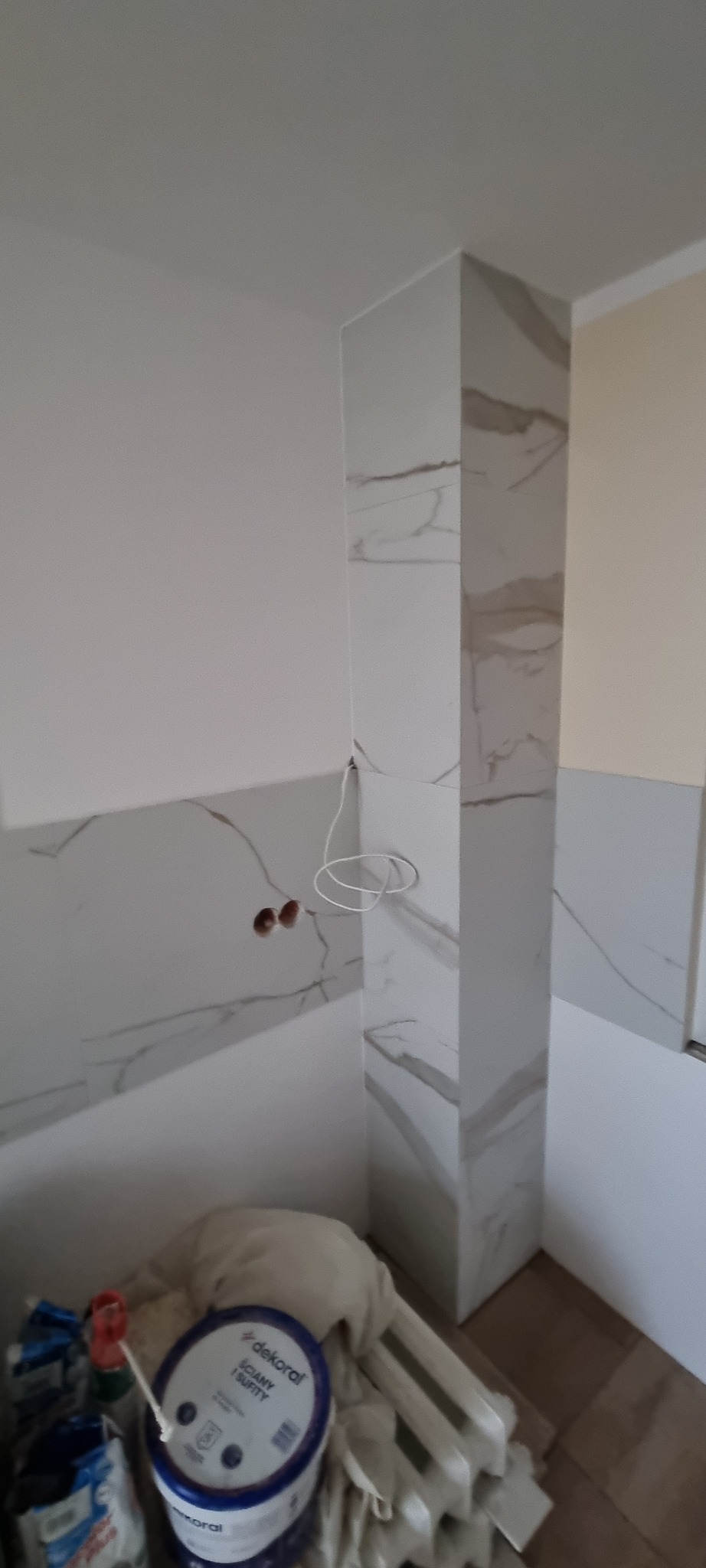 Fragment łazienki w trakcie remontu: częściowo położone płytki imitujące marmur na ścianach i pionowym słupie, widoczne puszki elektryczne i przewody, na podłodze wiadro z farbą do ścian i sufitów...