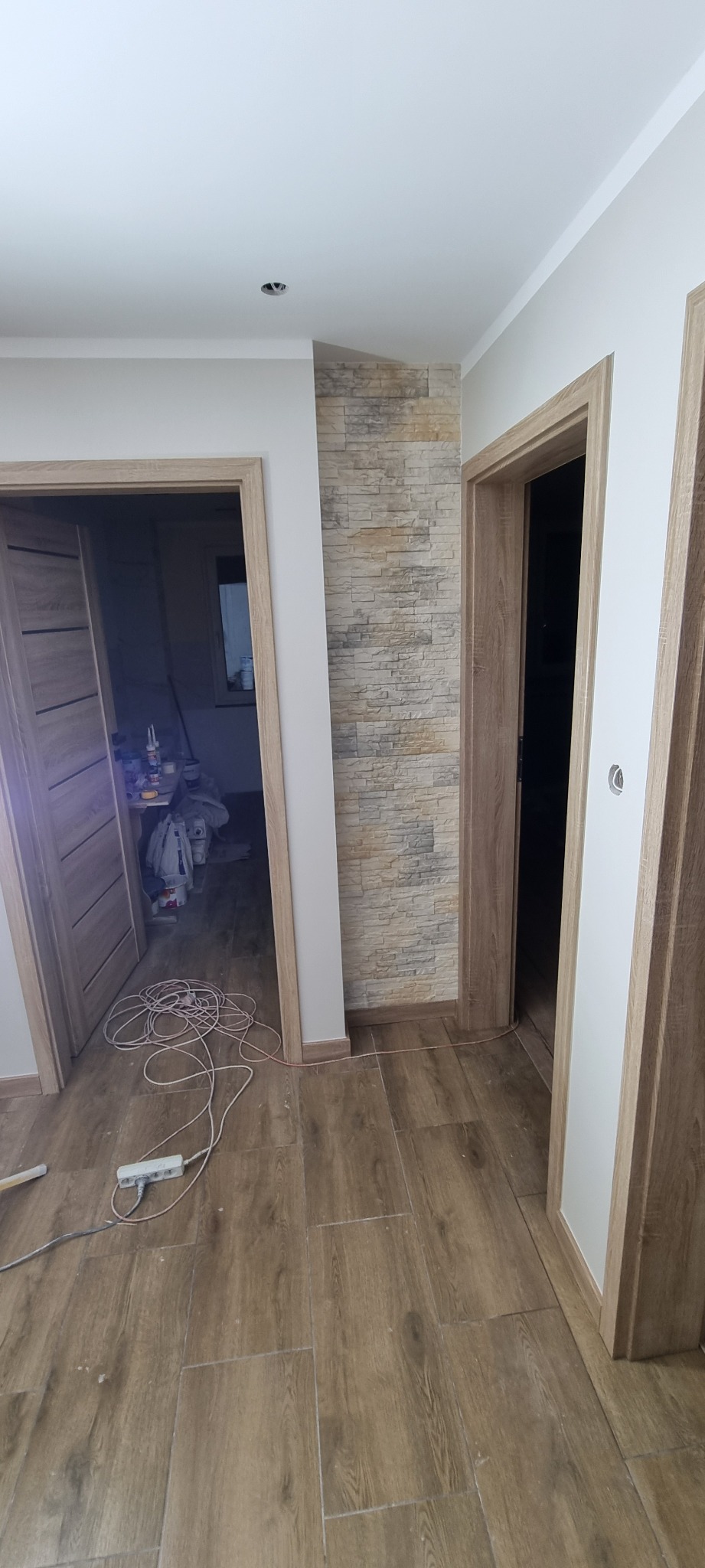 Wnętrze w trakcie remontu: położona podłoga z płytek imitujących drewno, widoczne trzy ościeżnice drzwiowe z zamontowanymi drzwiami po lewej, ściana wyłożona dekoracyjnym kamieniem, przedłużacz...