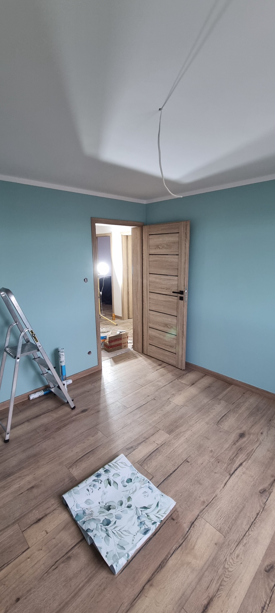 Pokój po malowaniu ścian na turkusowo, z drewnopodobną podłogą, otwartymi drzwiami i rozłożoną drabiną malarską.
