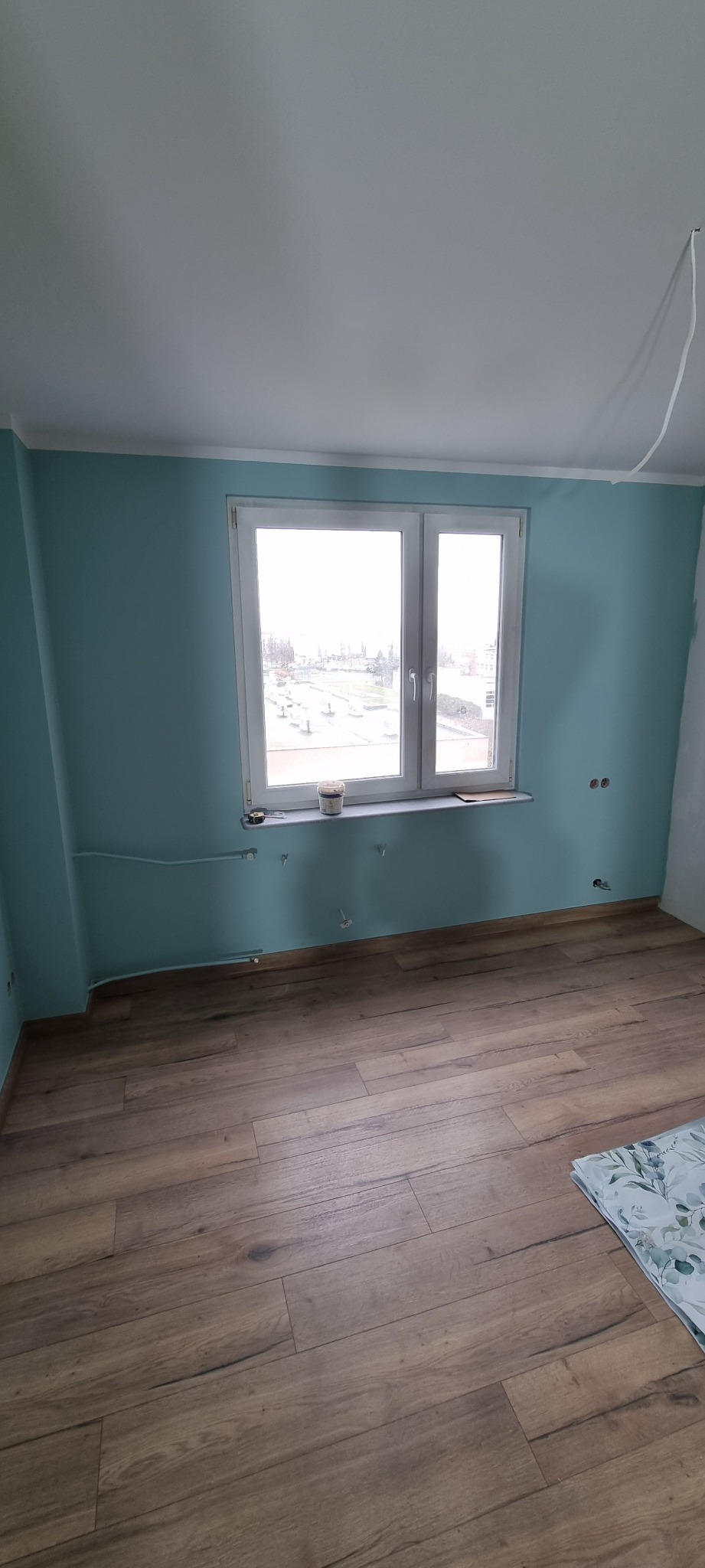 Pokój po remoncie z drewnopodobną podłogą, turkusowymi ścianami i białym sufitem; widoczne wystające kable i rury instalacji elektrycznej i hydraulicznej, oraz wiaderko z farbą i miarka...