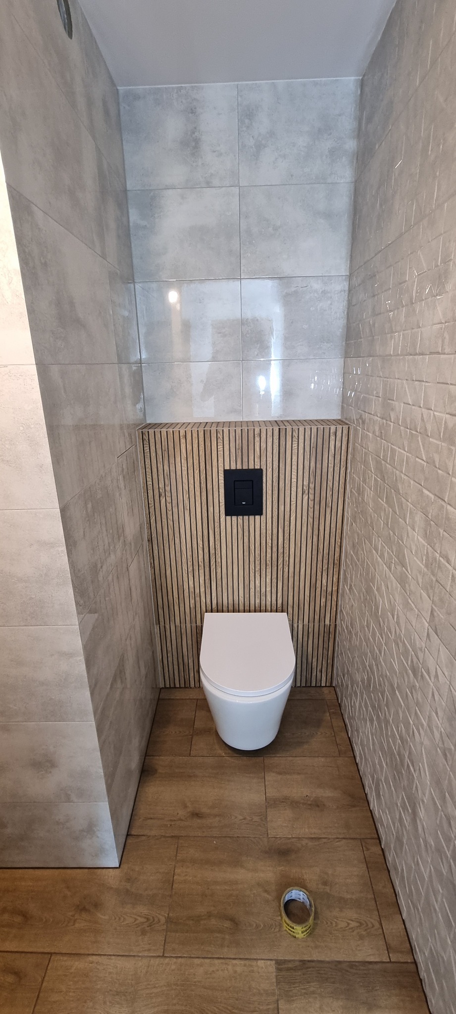 Nowoczesna toaleta podwieszana z czarnym przyciskiem spłukującym na tle drewnianych lameli, szara płytka imitująca beton na ścianach i podłodze, na podłodze leży taśma malarska.