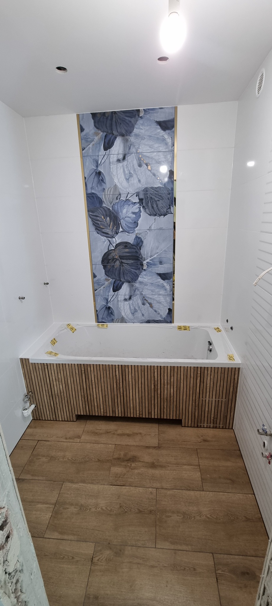 Nowoczesna łazienka z wanną obudowaną drewnianymi panelami, płytki z motywem roślinnym na ścianie, podłoga wyłożona panelami imitującymi drewno.