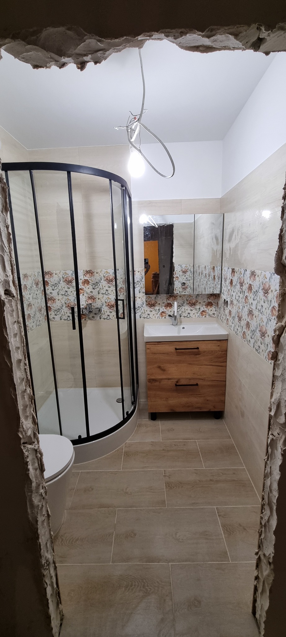 Niedokończona łazienka z kabiną prysznicową z czarnymi ramami, umywalką z szafką z drewnopodobnymi frontami i płytkami z motywem kwiatowym, widoczne surowe krawędzie ścian.