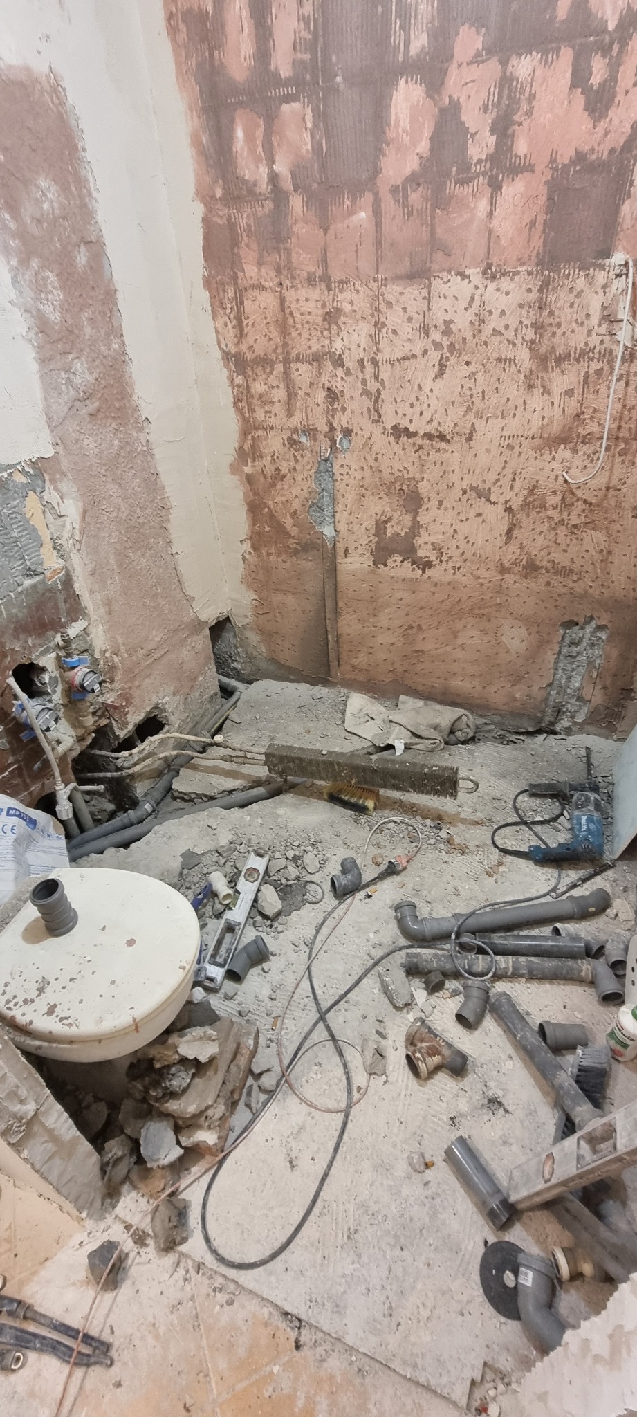 Demontaż instalacji wodno-kanalizacyjnej w trakcie remontu łazienki, widoczne rury, poziomica, wiertarka i gruz na podłodze, ściany częściowo obdarte z płytek i tynku.