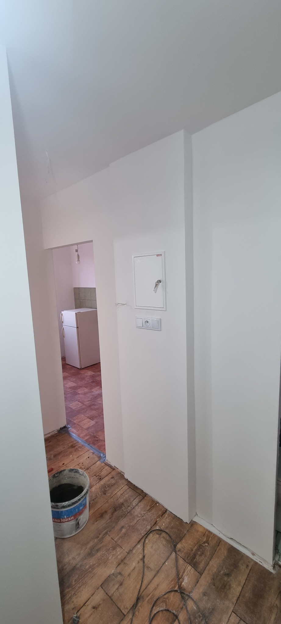 Świeżo pomalowane na biało ściany w mieszkaniu, widoczny fragment podłogi z drewnianych desek i wiadro z farbą. W tle widoczna kuchnia.