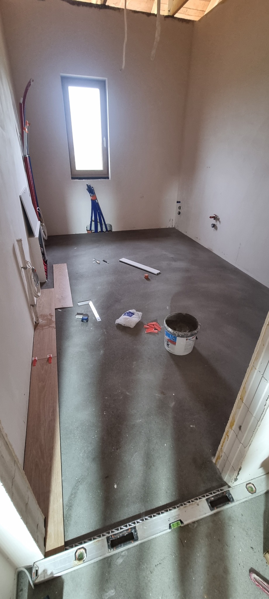 Wnętrze w trakcie prac: wylewka podłogowa, narzędzia glazurnicze, poziomica przy progu. Widoczne instalacje. Okno w tle.
