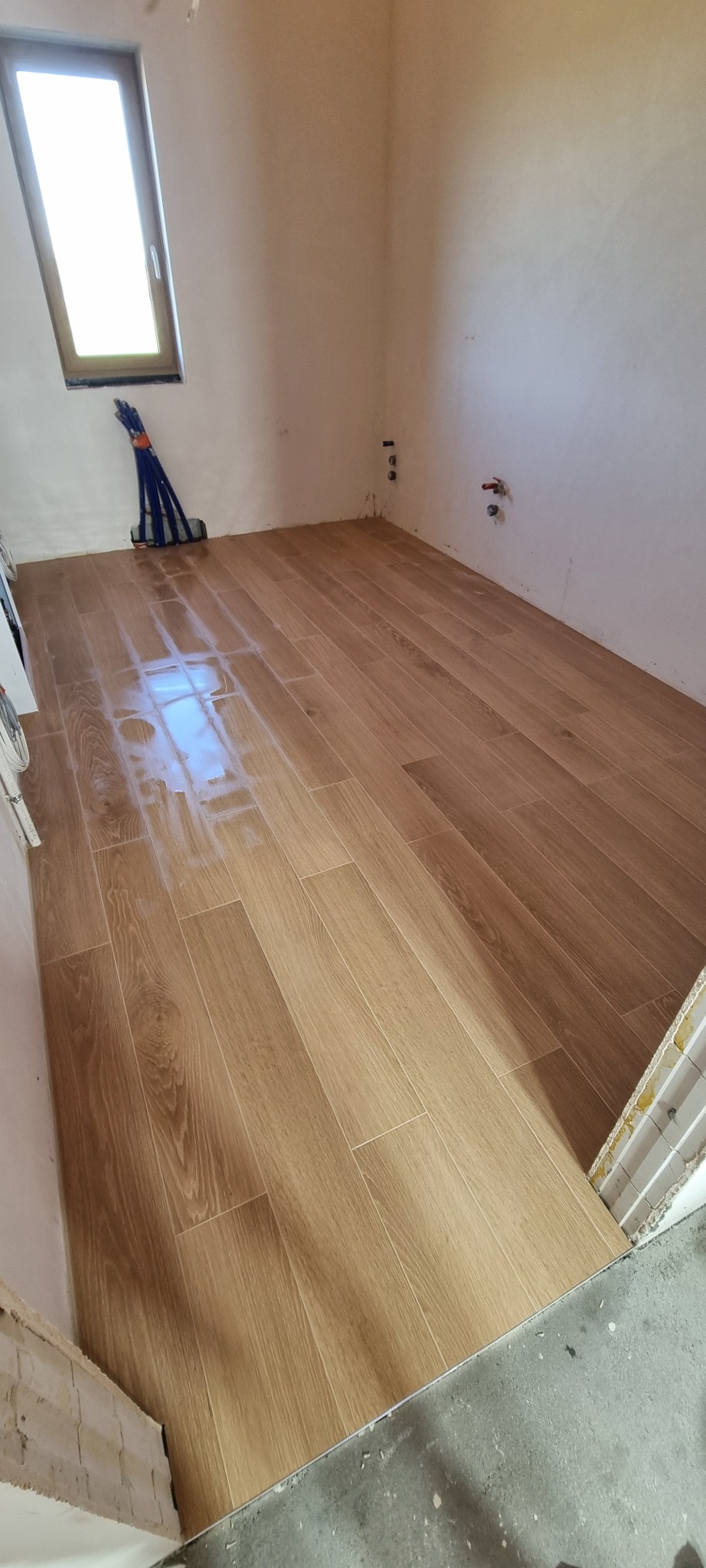 Ułożone, podłużne płytki podłogowe imitujące drewno w jasnym odcieniu, widok z góry, fragment ściany z oknem i nieotynkowane fragmenty ścian.