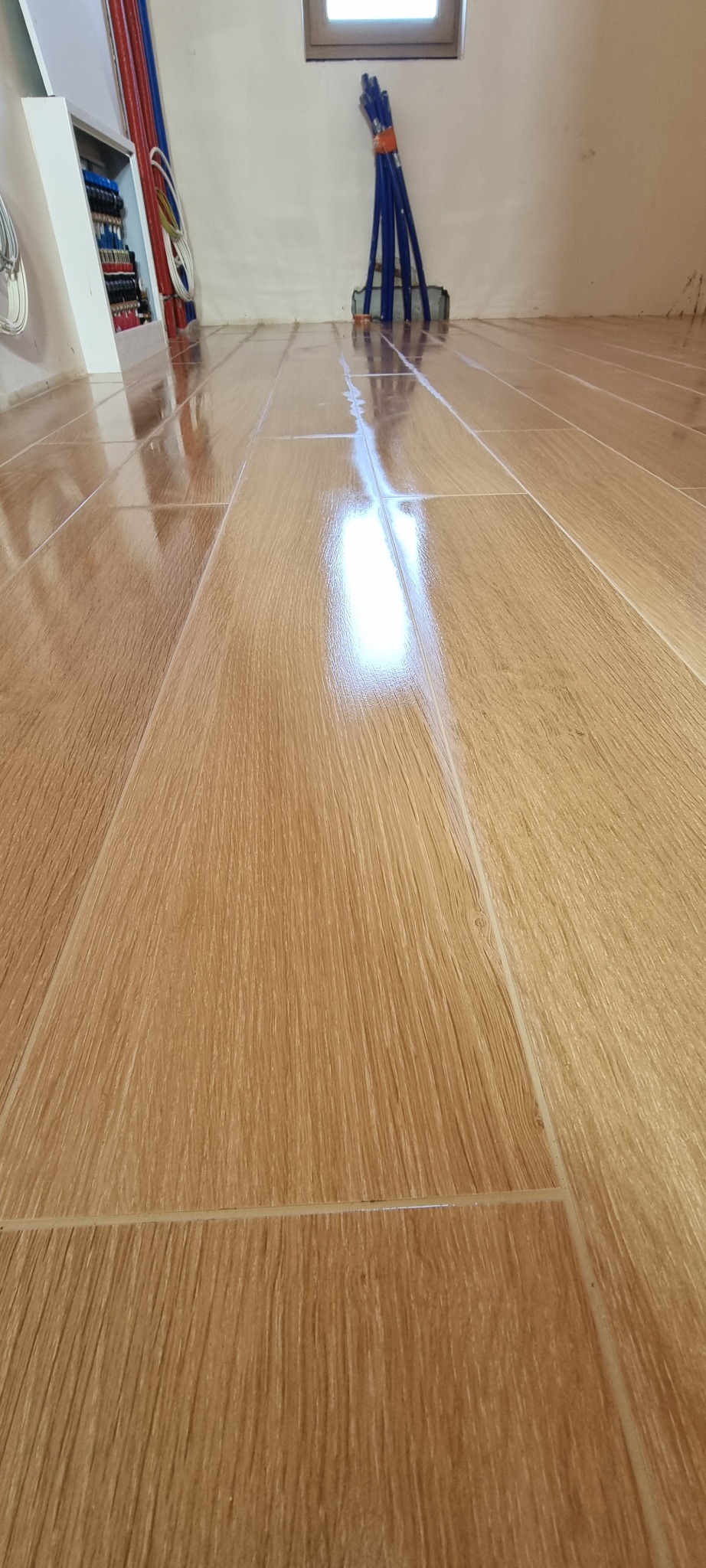 Połyskujące, drewnopodobne płytki na podłodze w nowym wnętrzu. Widoczne fragmenty instalacji grzewczej i okno w tle. Ujęcie z niskiej perspektywy, podkreślające fakturę.