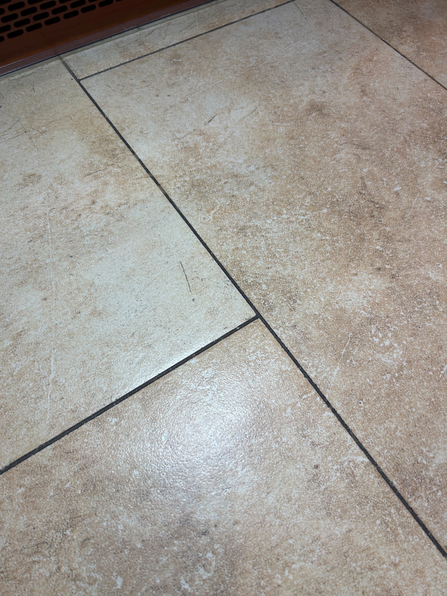 Beżowe płytki podłogowe z ciemnymi fugami, widoczne zabrudzenia i drobne rysy na powierzchni, fragment kratki wentylacyjnej w górnej części kadru.