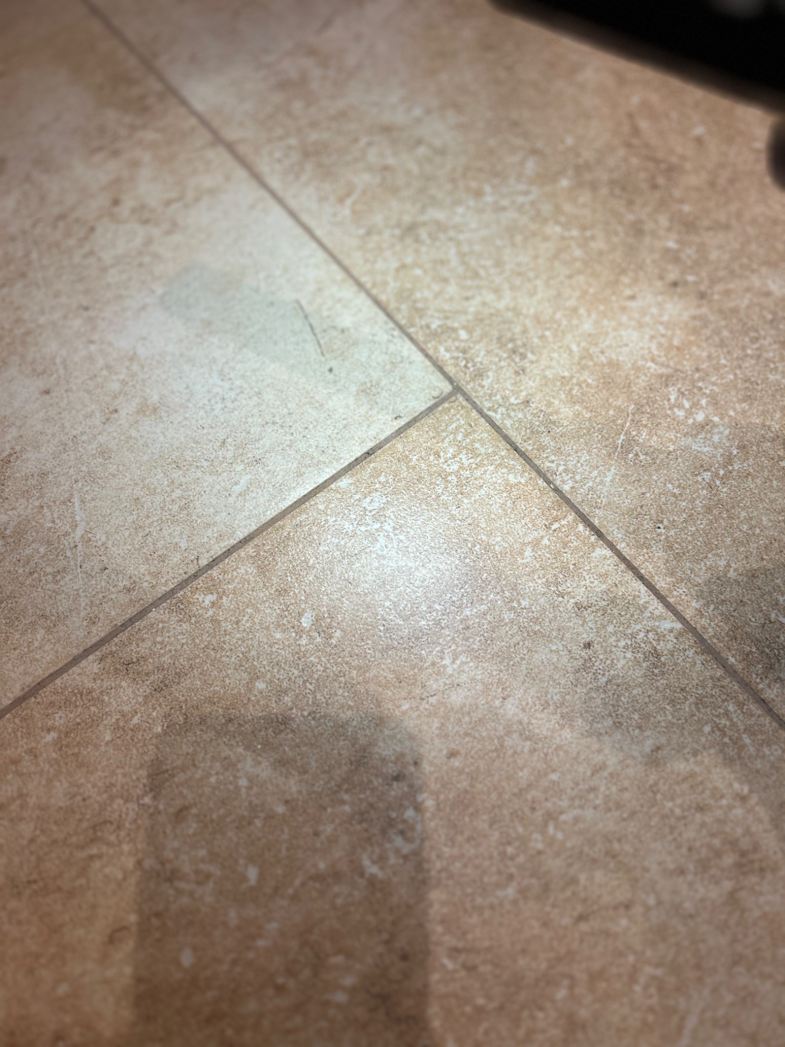 Beżowe płytki podłogowe z delikatną strukturą, ułożone w kwadraty, widoczne fugi, z fragmentem zacienionego obiektu w rogu.