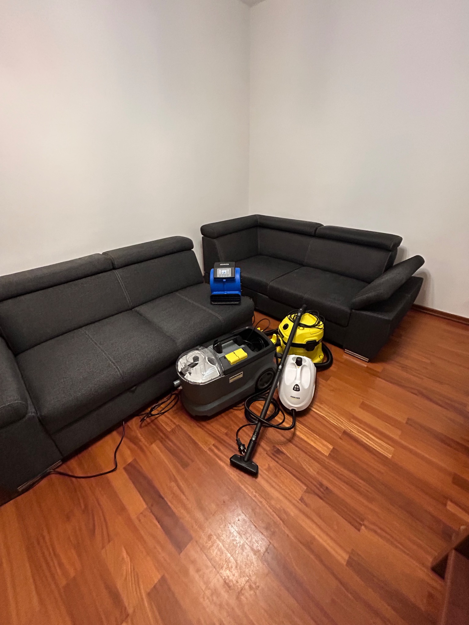 Dwie szare sofy narożne w pokoju z parkietem, obok nich sprzęt do czyszczenia tapicerki: odkurzacz piorący Kärcher, parownica i mały odkurzacz do mierzenia wilgotności.