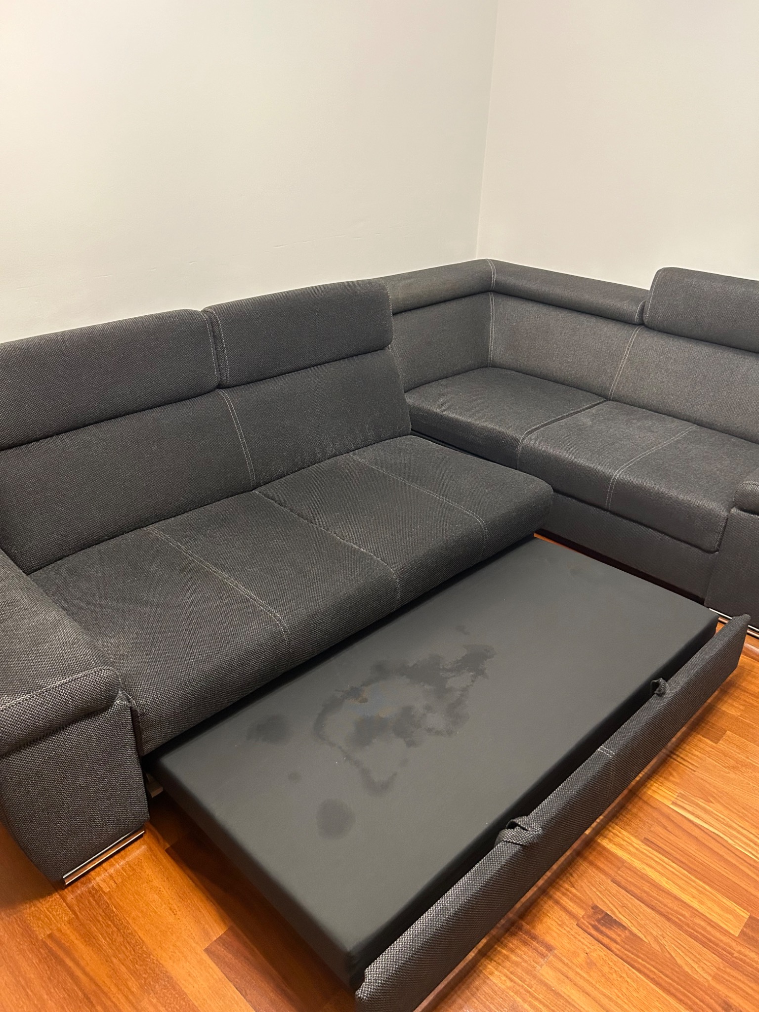 Rozłożony narożnik z szarej tkaniny z widocznymi plamami na wysuniętym elemencie do spania, na parkiecie.