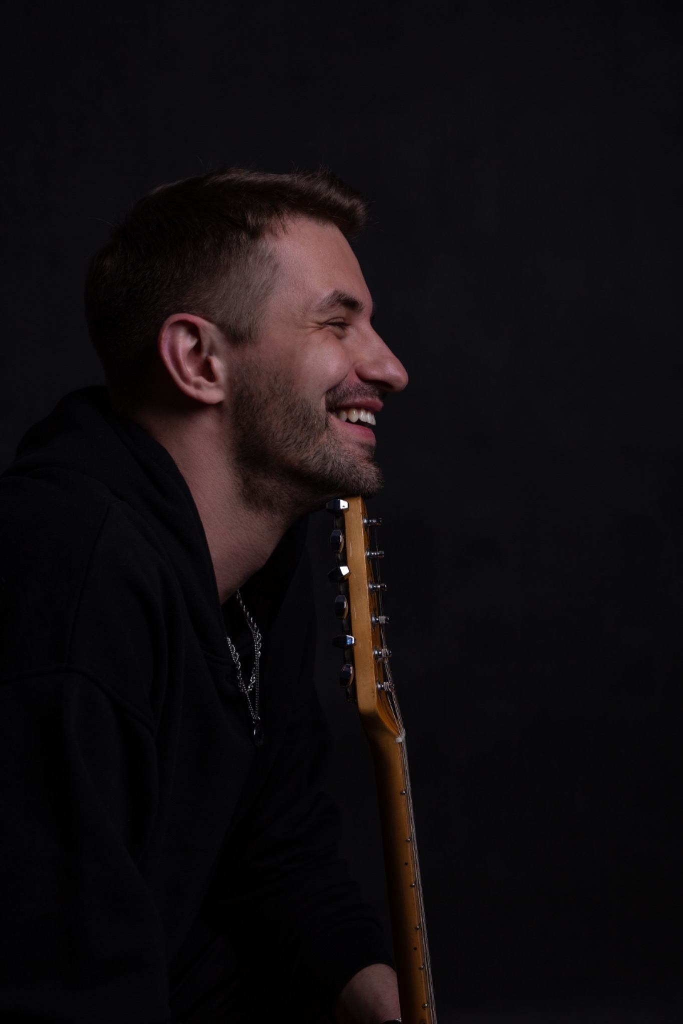 Uśmiechnięty mężczyzna z brodą, profil, opierający szyję o gryf gitary elektrycznej, ubrany w czarną bluzę z kapturem i srebrny łańcuszek na ciemnym tle.
