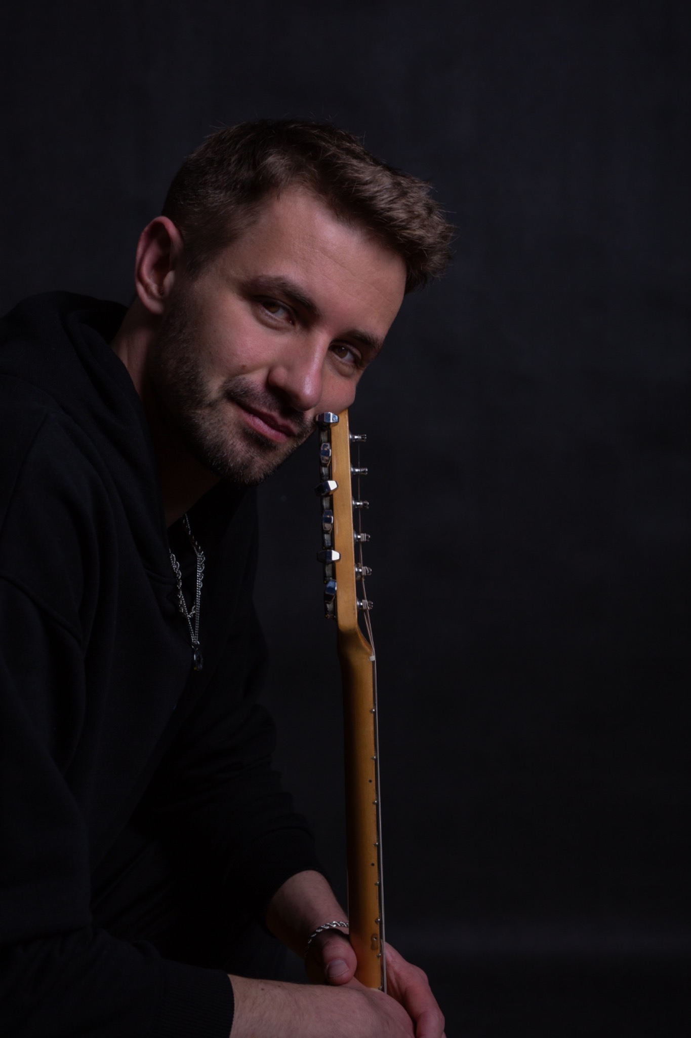 Portret mężczyzny z gitarą elektryczną na ciemnym tle, ubrany w czarną bluzę z kapturem, z delikatnym uśmiechem i srebrnym łańcuszkiem.