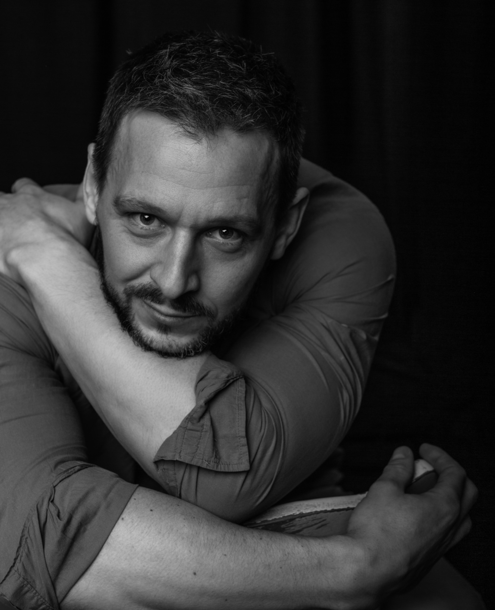 Portret biznesowy mężczyzny z brodą, w koszuli, obejmującego się ramionami, na czarnym tle.