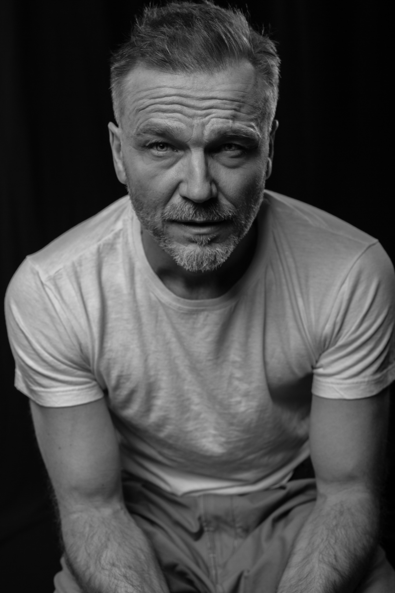 Czarno-białe portretowe ujęcie dojrzałego mężczyzny z krótkimi, siwiejącymi włosami i brodą, w białym T-shircie, na ciemnym tle.