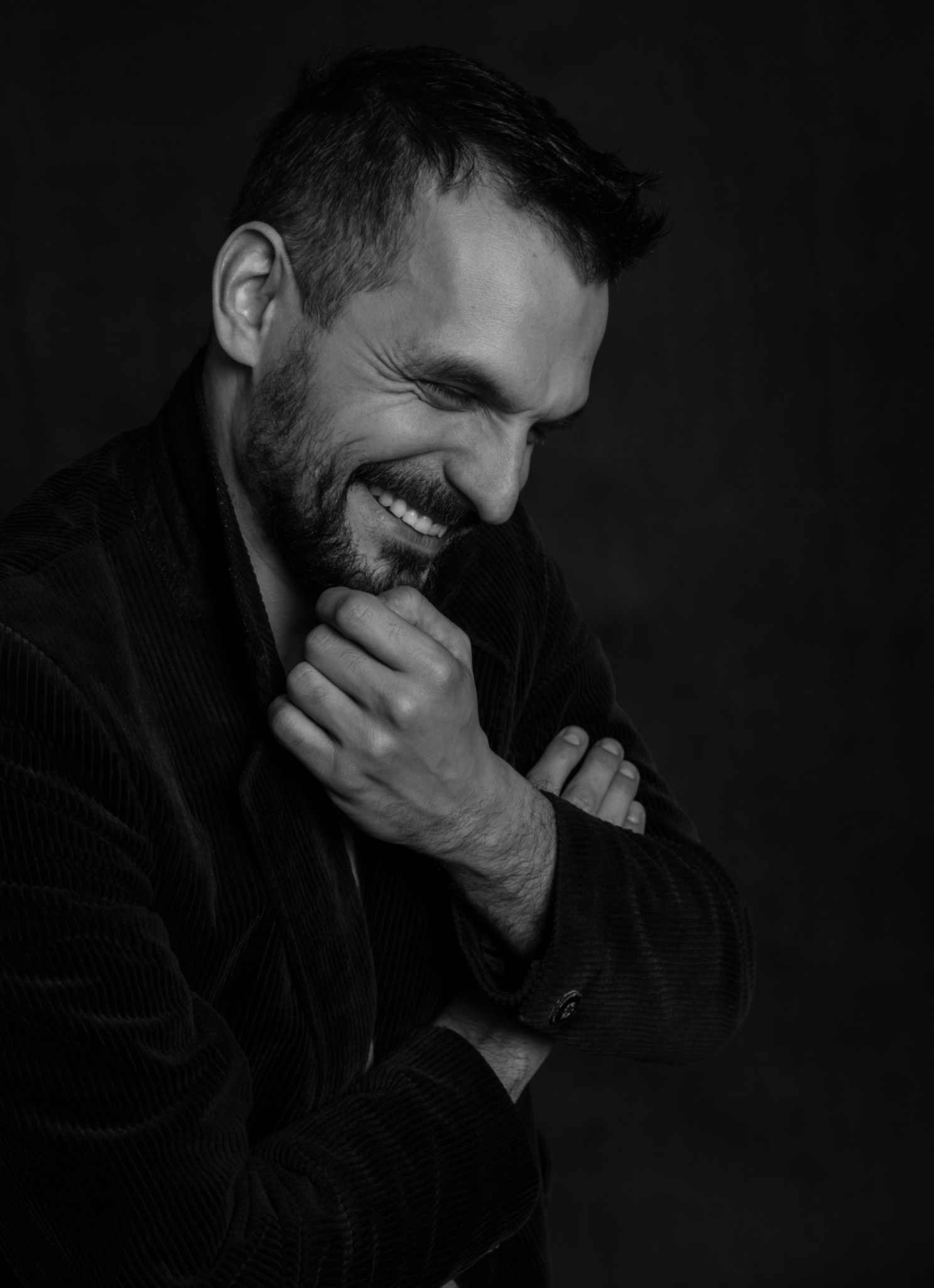 Czarno-białe portretowe zdjęcie uśmiechniętego mężczyzny z brodą, w ciemnej, aksamitnej marynarce, z rękami skrzyżowanymi i dłonią dotykającą brody, na ciemnym tle.