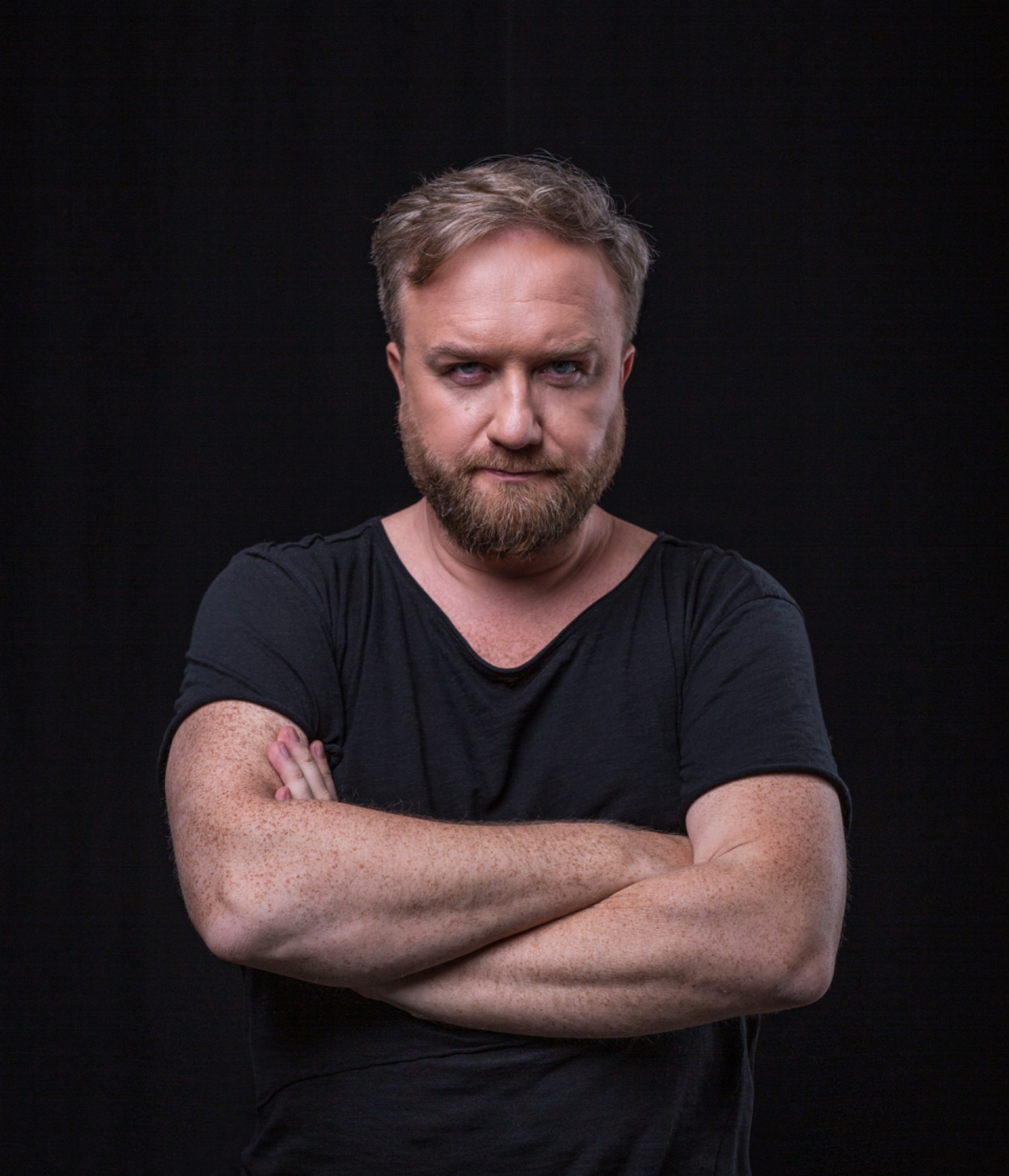 Portret mężczyzny z brodą i piegami, na czarnym tle, z założonymi rękami i intensywnym spojrzeniem.