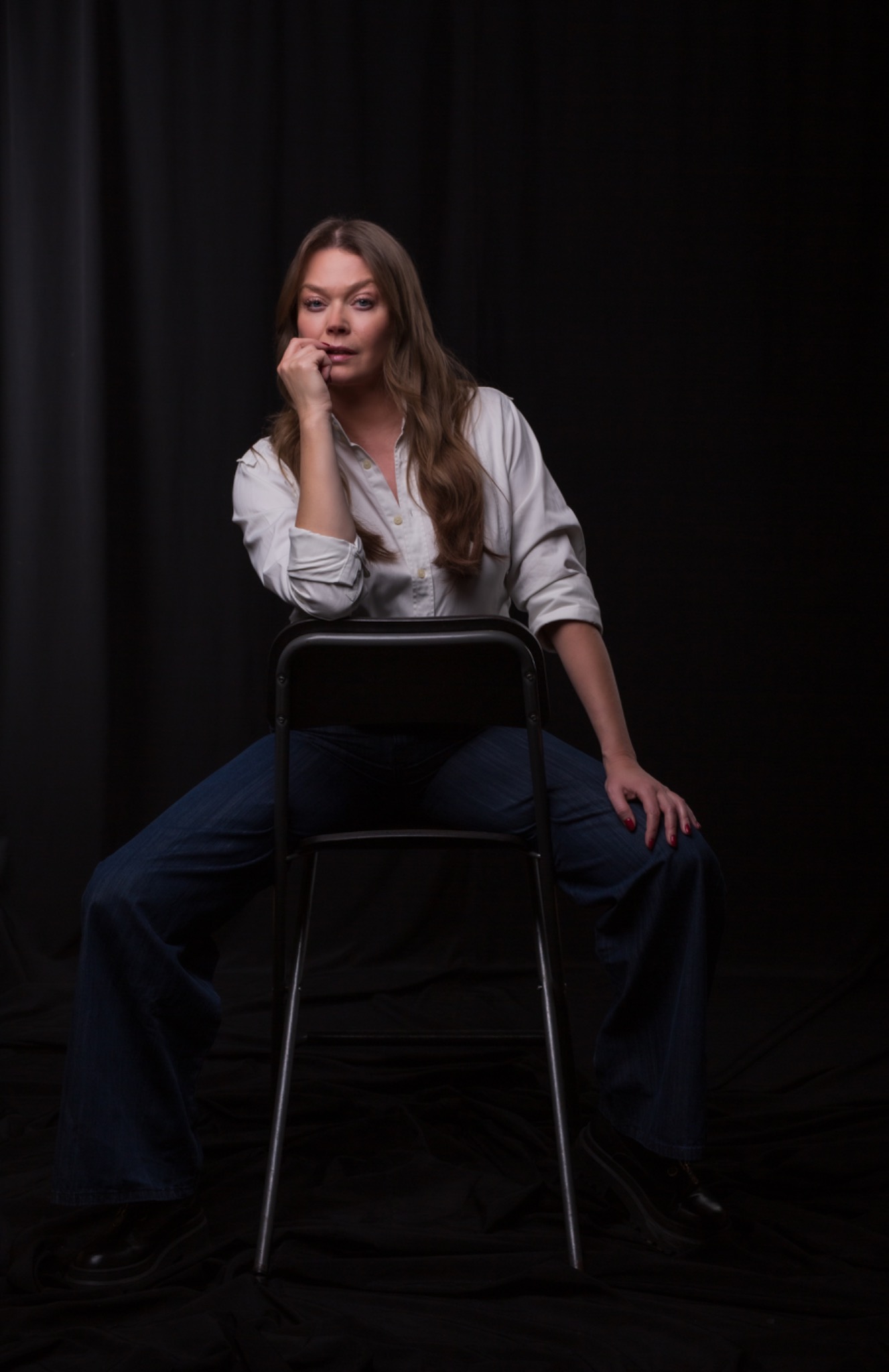 Kobieta w białej koszuli i dżinsach siedzi na metalowym krześle na czarnym tle, zamyślona, z ręką przy ustach.