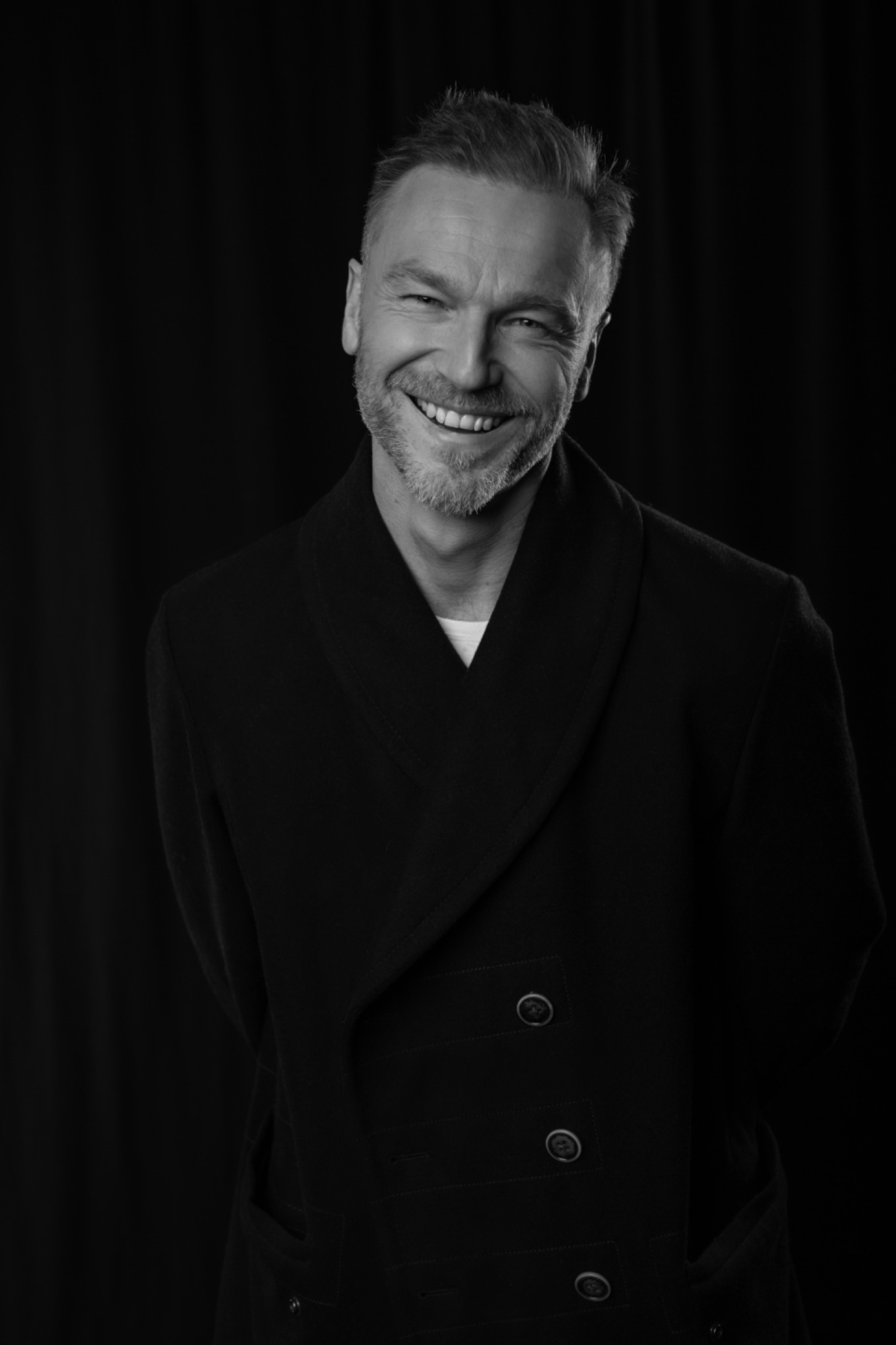 Czarno-białe portretowe ujęcie uśmiechniętego mężczyzny z siwiejącą brodą, ubranego w ciemny płaszcz na tle czarnego materiału.