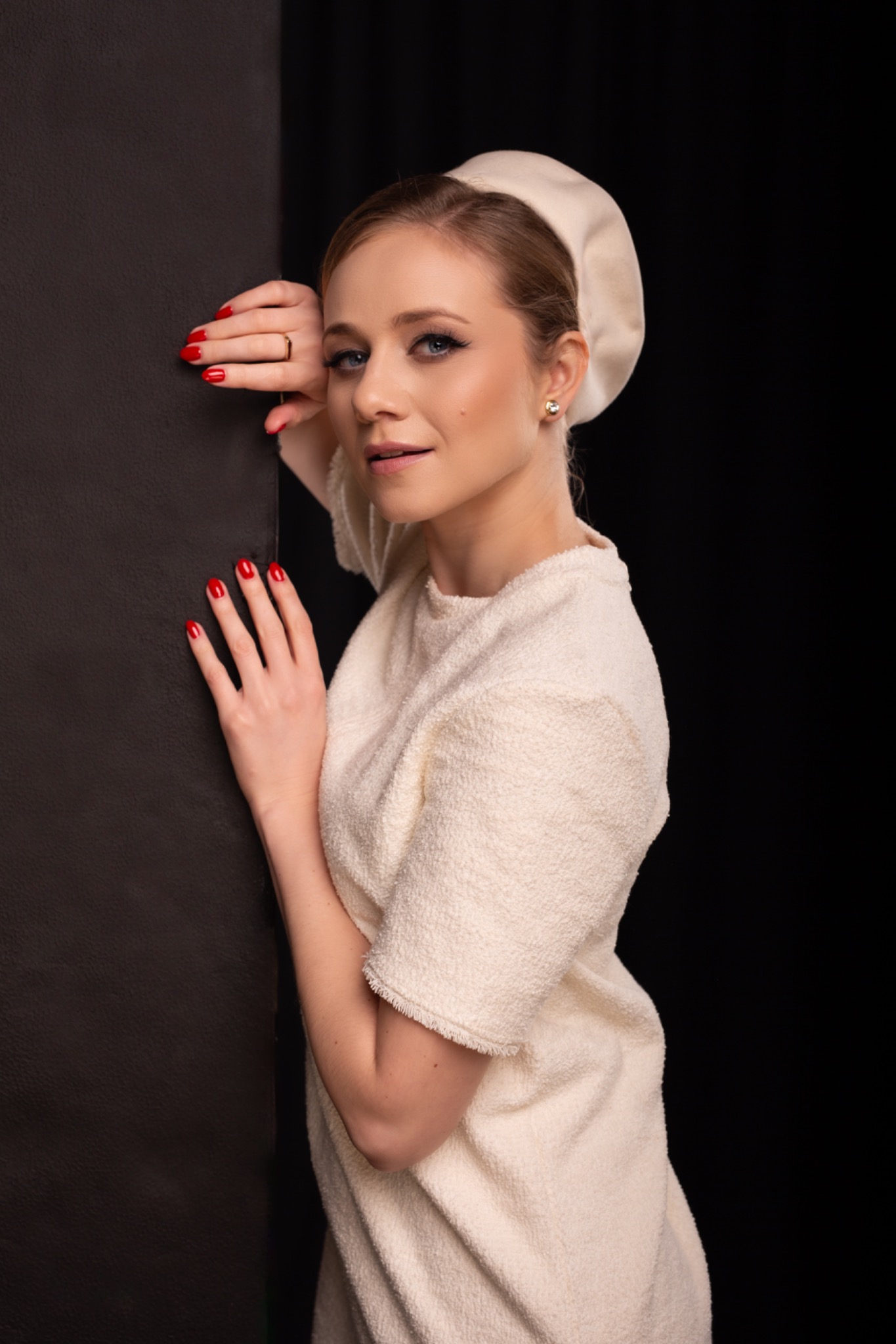 Portret kobiety w kremowej sukience i beretce, z czerwonym manicure, opierającej się o ciemną ścianę na czarnym tle.