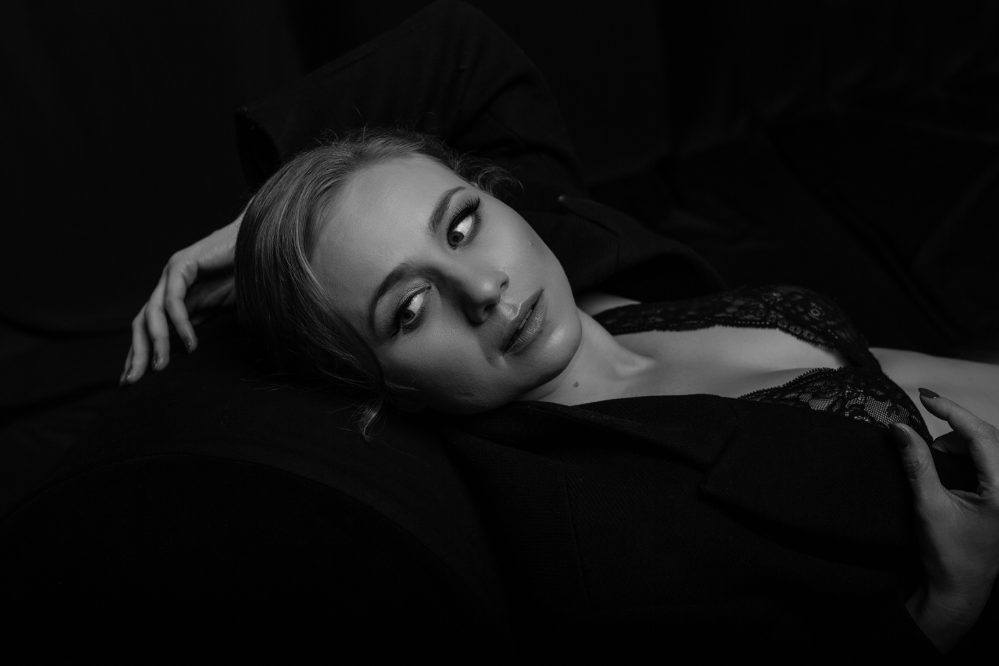 Czarno-białe portretowe ujęcie kobiety w bieliźnie i marynarce, leżącej na ciemnym tle, wykonane w Krakowie, z delikatnym oświetleniem podkreślającym jej rysy twarzy.