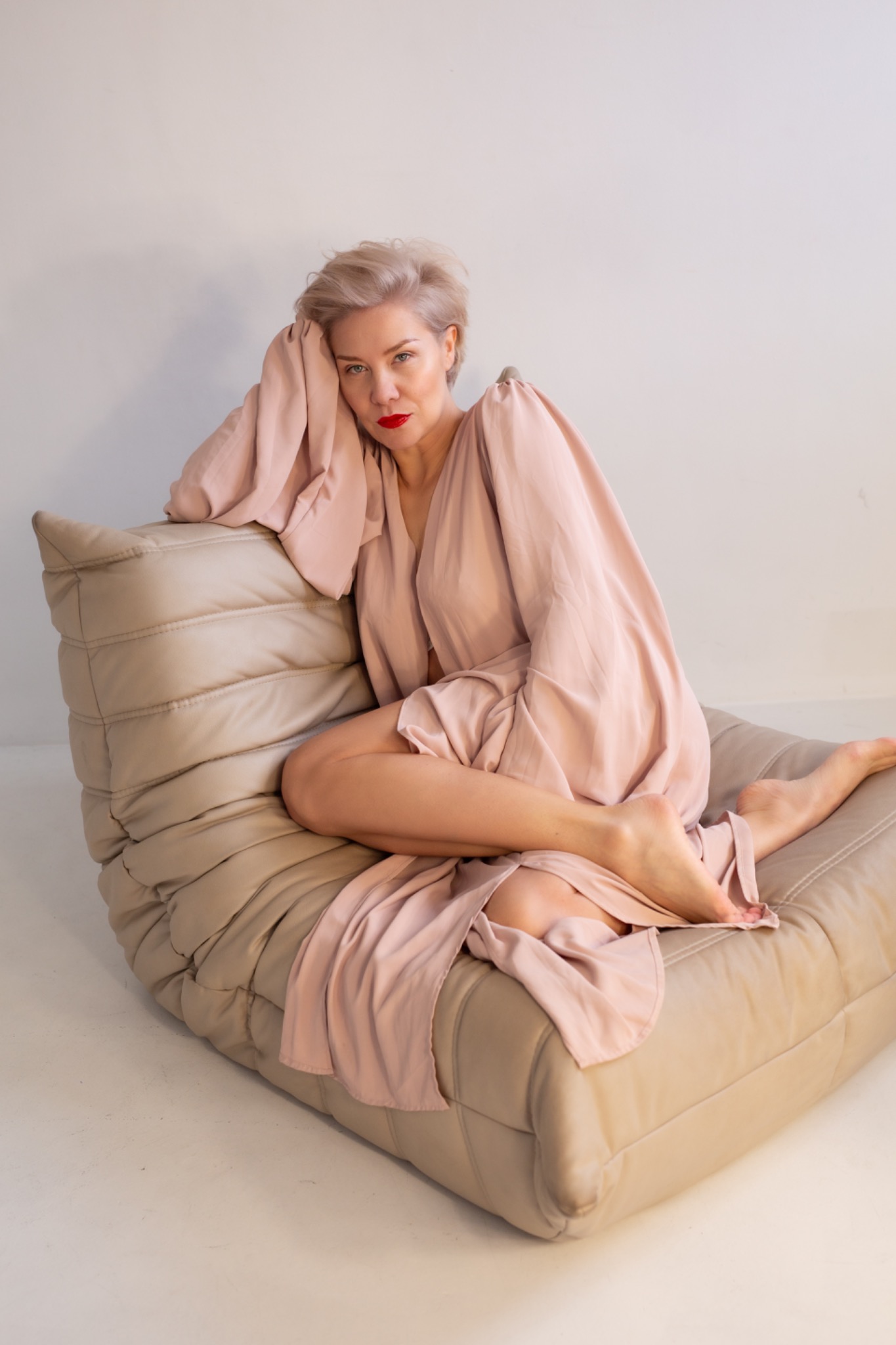 Kobieta z krótkimi blond włosami i czerwoną szminką pozuje w luźnej, pudrowej sukience na beżowej, skórzanej pufie na tle jasnej ściany. Uwagę przyciąga wyrazisty makijaż.