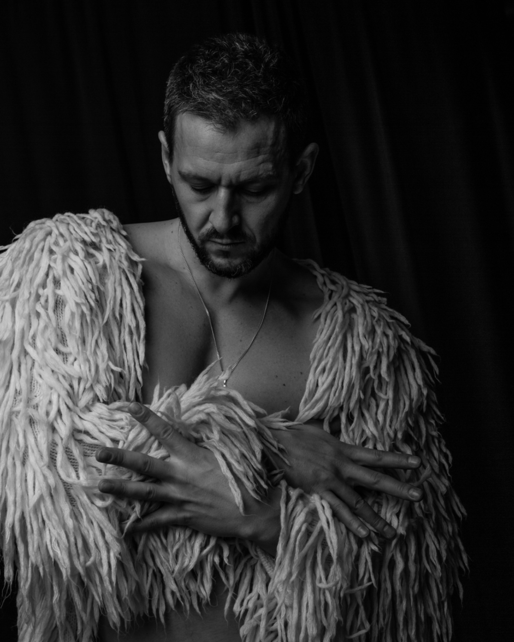 Czarno-białe zdjęcie portretowe mężczyzny z brodą, spoglądającego w dół, ubranego w futrzaną narzutkę z długich, grubych pasm, z rękami skrzyżowanymi na klatce piersiowej i delikatnie widocznym...