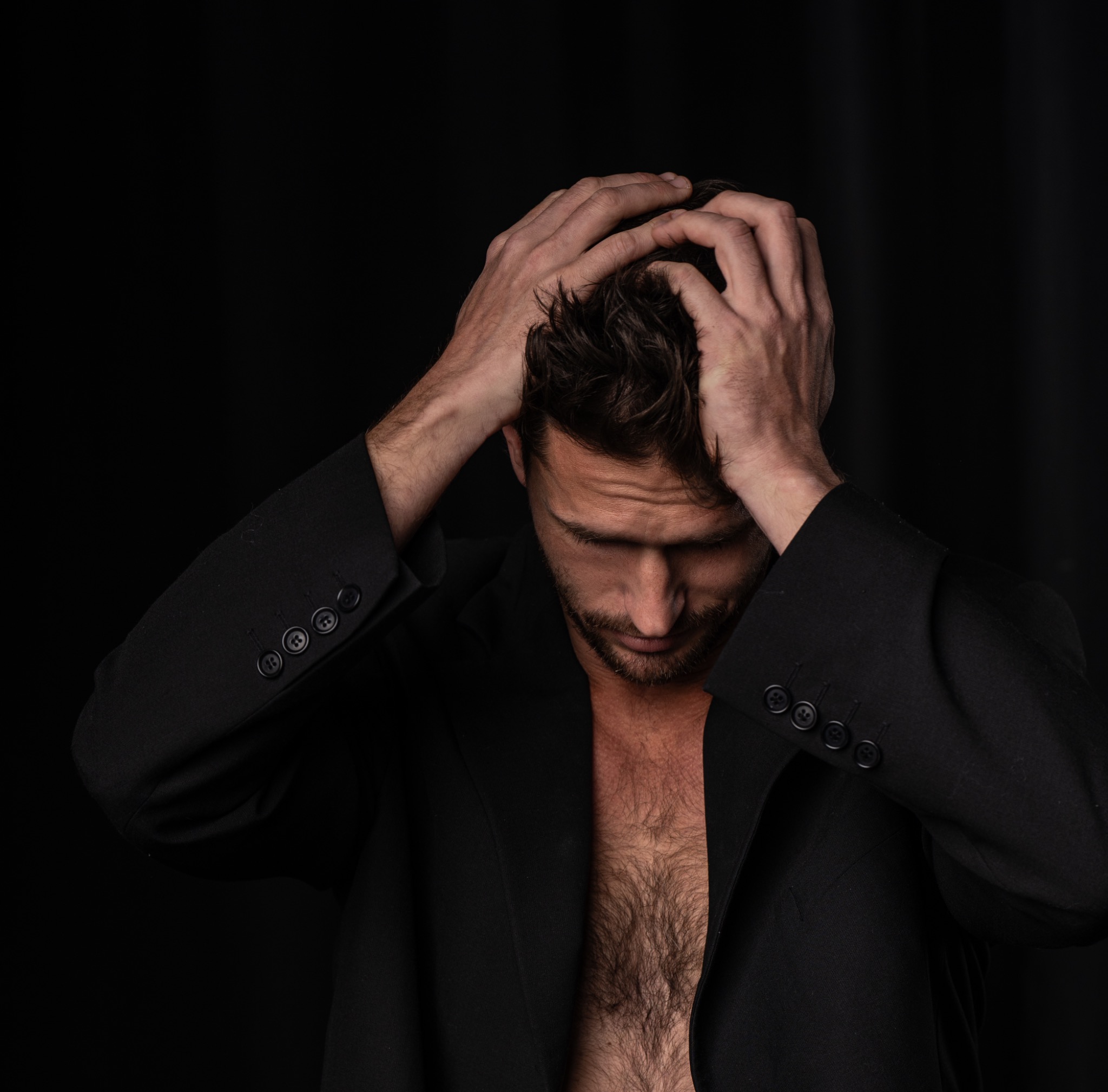 Mężczyzna w czarnej marynarce z rozpiętą koszulą, zamyślony, dłońmi dotyka włosów na ciemnym tle.